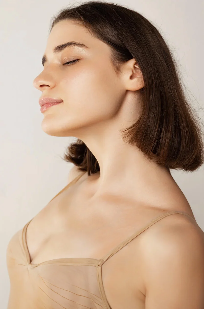 Close-up profile of a woman with short, dark brown hair and closed eyes, exuding serenity. Her skin is smooth and flawless, and she wears a light beige, strapless or spaghetti strap bra with a delicate, textured fabric. The background is a soft, neutral tone, enhancing the soft lighting that highlights her features and skin. - Acne Scar Removal in Los Angeles, CA