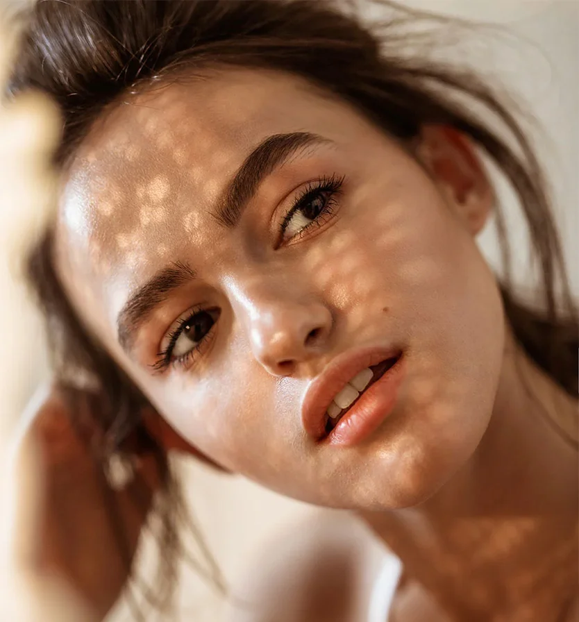 Close-up portrait of a young woman with light brown hair pulled back. Her smooth, glowing skin is illuminated by sunlight, creating patterns of light and shadow on her face. She has dark brown eyes, well-defined eyebrows, and a neutral expression with slightly parted lips. The blurred beige or cream background keeps the focus on her face. - Acne Scar Removal in Los Angeles, CA
