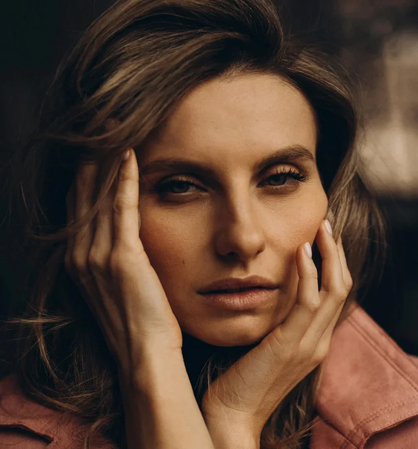 A close-up portrait of a woman with long, light brown waves framing her face. Her expression is thoughtful, with her gaze slightly downward and hands resting gently on her temples. She wears light makeup and a soft brownish-pink jacket. The dark, blurred background contrasts with her illuminated face, emphasizing her features with subtle highlights and shadows. - BBL Laser in Los Angeles, CA