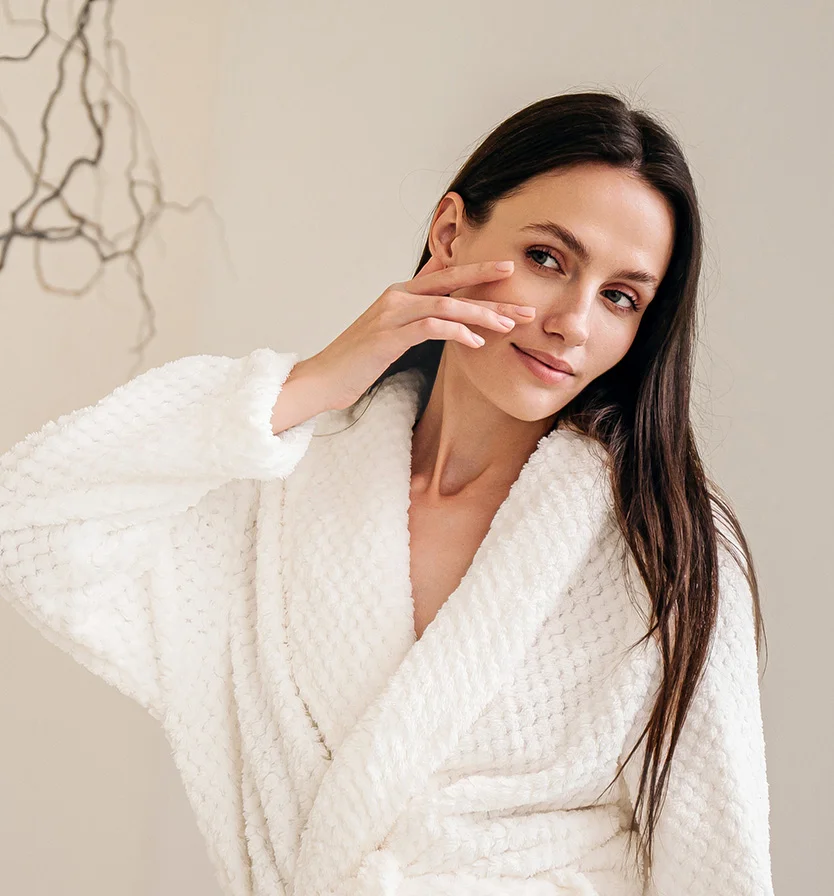 A woman with long, dark brown hair and a light complexion is seen from mid-chest up, wearing a textured, cream-colored bathrobe. Her neutral expression is soft, with her gaze slightly averted. One hand rests gently on her cheek. The light beige background is minimal, with a faint silhouette of a branch. - Chemical Peels in Los Angeles, CA