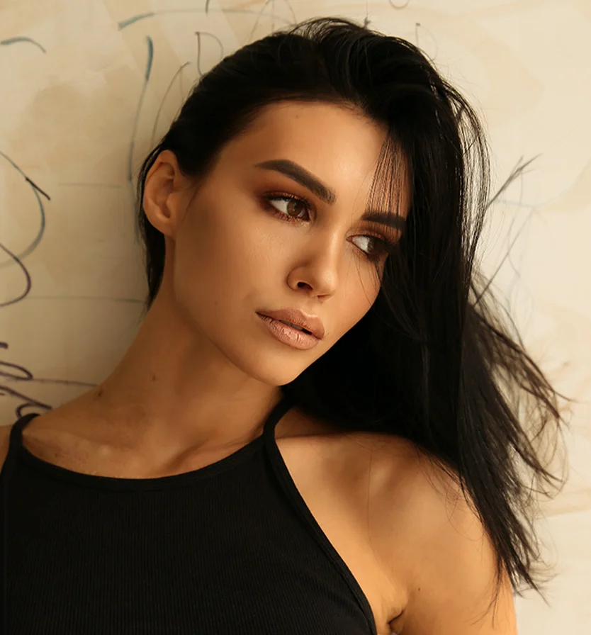 A close-up portrait of a woman with long, tousled dark hair framing her face. She has a light tan complexion, well-defined features, light brown eyes, and dark brown eyebrows. Her lips are a soft beige or peachy nude. She wears a black ribbed tank top with thin straps. The light beige background has faint, indistinct markings. Soft lighting enhances her features, creating a natural beauty portrait. - Dermal Fillers in Los Angeles, CA