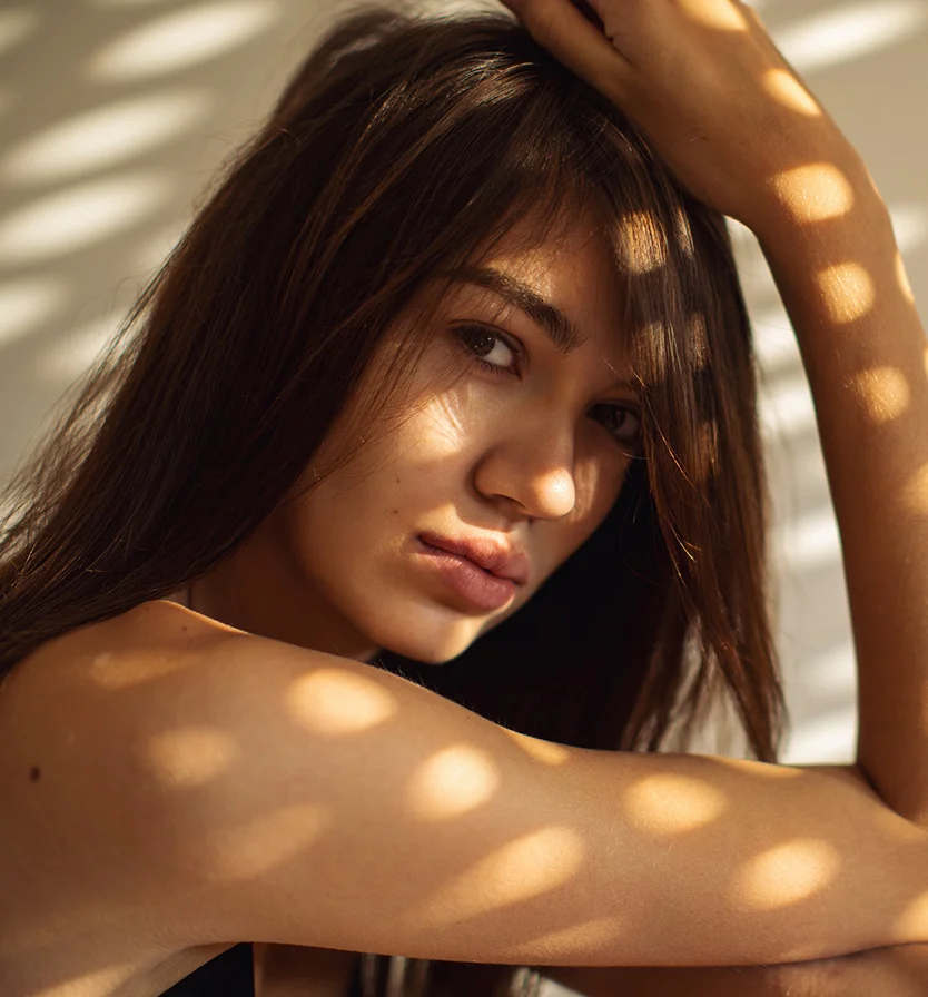 A young woman with long brown hair gazes slightly away from the camera. Soft, dappled light casts patterns on her face and arms, highlighting her smooth skin and light brown eyes. The focus is on her thoughtful expression and the play of light and shadow. - Facials in Los Angeles, CA