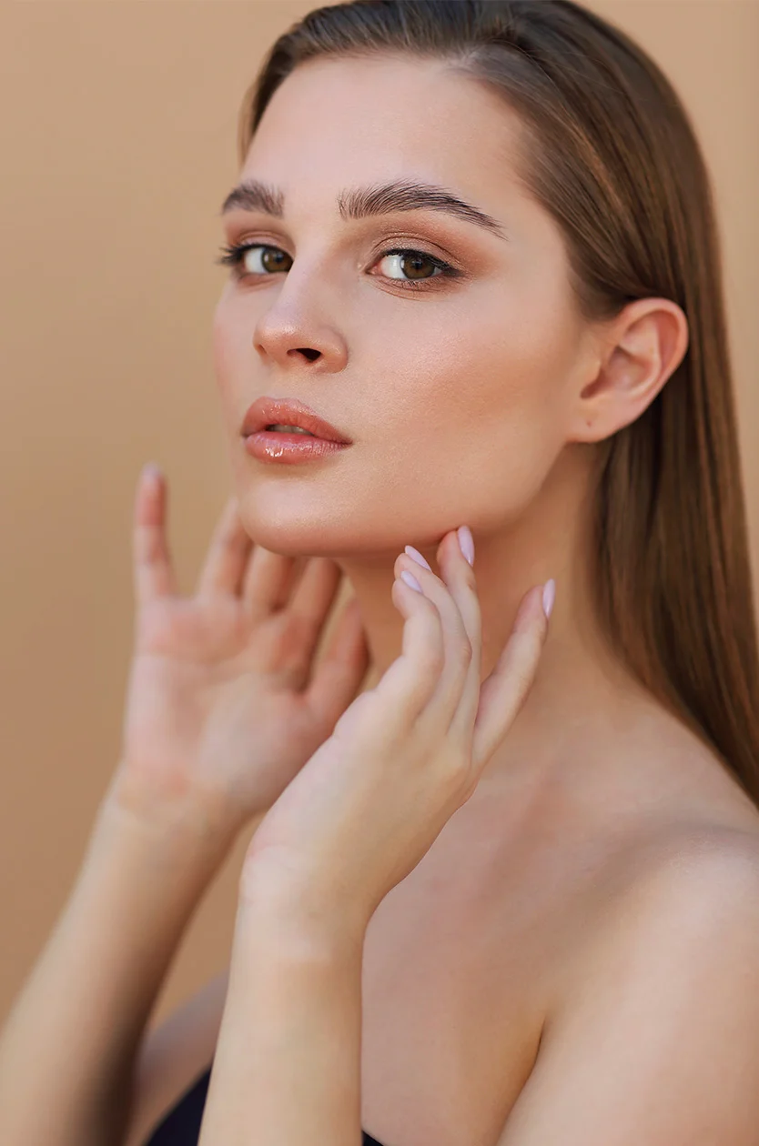 A young woman with smooth, light-toned skin and long brown hair has a serene expression. She wears subtle makeup and gently rests her hands on her face and neck. The background is muted beige, with soft, even lighting highlighting her features. - Hydrafacial in Los Angeles, CA