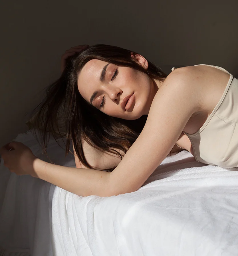 A woman lies on a white bedsheet, resting her head on one arm while the other drapes down her side. Her eyes are closed, with a relaxed expression, and her dark hair flows around her face. She wears a light beige tank top, with soft light casting subtle shadows on her body. The textured sheet provides a neutral backdrop, keeping the focus on her serene appearance. - Hyperhidrosis in Los Angeles, CA