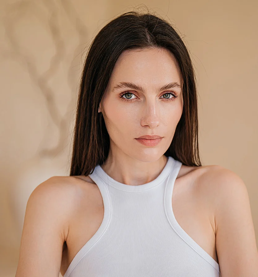 Close-up of a woman with long, dark brown hair and a light complexion. She wears a white ribbed tank top, with a neutral expression and light brown or hazel eyes. Her subtle makeup includes rosy blush and light lipstick. The background is beige or light brown, with a slight blurred shape, while even lighting highlights her face and shoulders. - IPL Photofacial in Los Angeles, CA