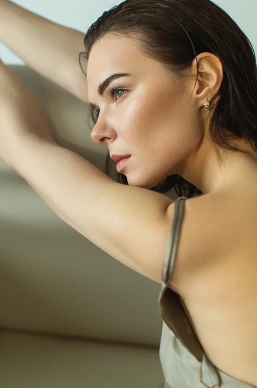 A woman with wet, dark brown hair wears a light taupe sleeveless top, her expression calm and reflective. Soft lighting highlights her glowing skin, and the blurred background emphasizes her serene presence. - Laser Facial in Los Angeles, CA
