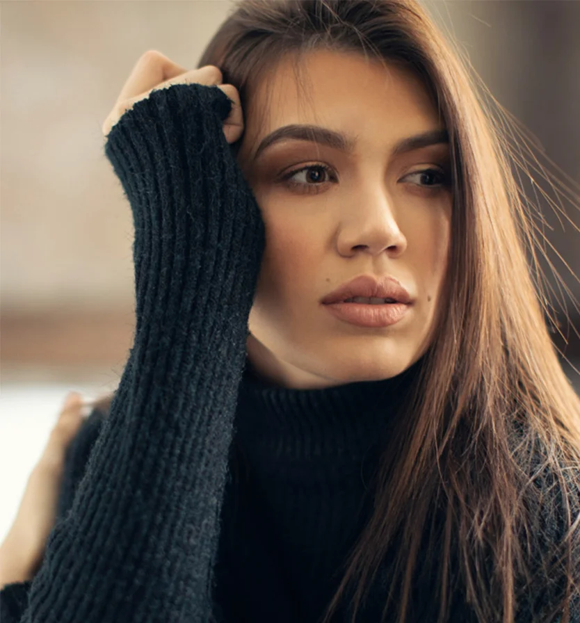 A young woman with long brown hair and a pensive expression wears a dark gray turtleneck sweater with textured knit. Her hand rests on her forehead as she gazes slightly to the right. Subtle makeup enhances her features, with natural light softly illuminating her face. - Laser Facial in Los Angeles, CA