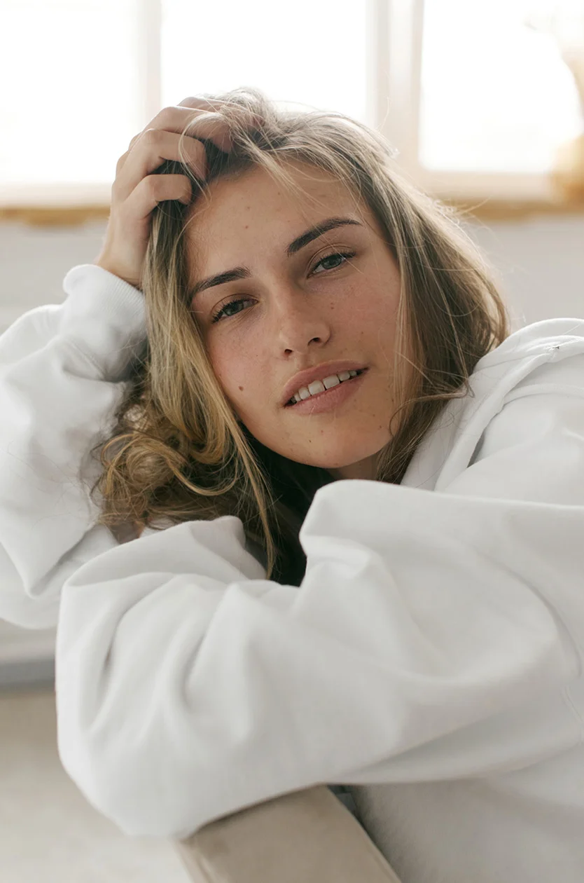 Close-up of a light-skinned young woman with shoulder-length, slightly wavy blonde hair. Her gaze is soft and contemplative, directed slightly to her right. She wears a white, long-sleeved sweatshirt with loose-fitting sleeves. The background is a light beige, possibly a couch or similar seating, with a soft, diffused light creating a relaxed, calm atmosphere. The focus is on her face and upper torso. - Laser Skin Resurfacing in Los Angeles, CA