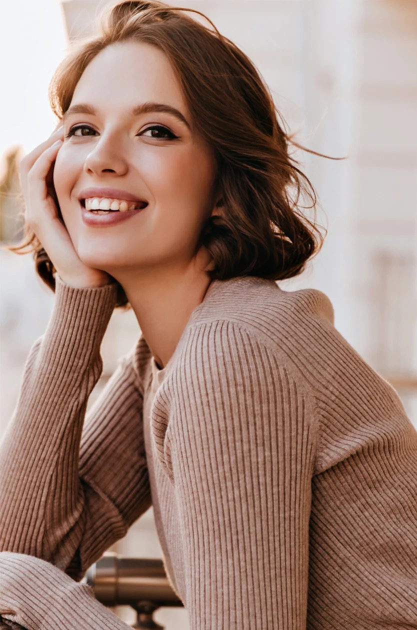 A young woman with light brown/auburn hair styled in a tousled bob smiles warmly at the camera. Her complexion is natural, with medium brown eyes. She wears a well-fitting light taupe ribbed knit sweater and gently places her hands on her face and neck. The background is blurred, hinting at an outdoor setting, with soft, diffused light illuminating her face, creating a bright and pleasant tone. - Microneedling with PRP RF in Los Angeles, CA