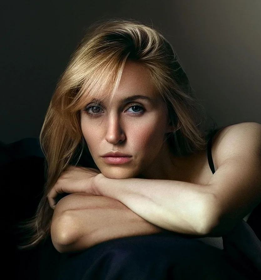 A woman with shoulder-length light brown hair gazes downward, her expression thoughtful. She has a light complexion and grayish-blue eyes. Resting her head and elbows on a surface, she wears a sleeveless black top. The soft, diffused lighting highlights her features and creates subtle shadows, with a dark, neutral background that emphasizes her face. - Microneedling with PRP RF in Los Angeles, CA